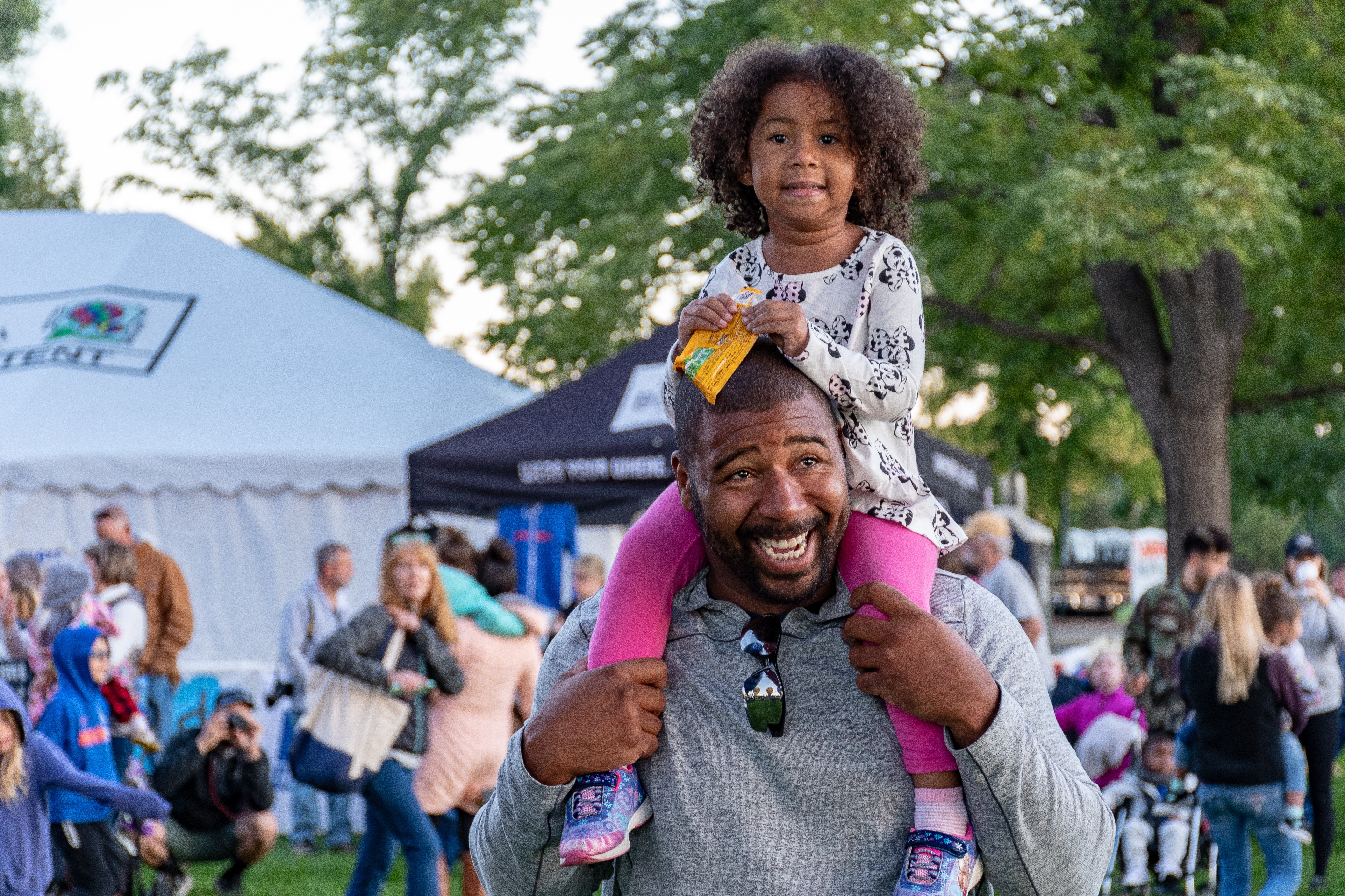 Man giving his daughter a ride on his shoulders, He is smiling and looking away from the camera. She is smilimg and lookimg into the camera.