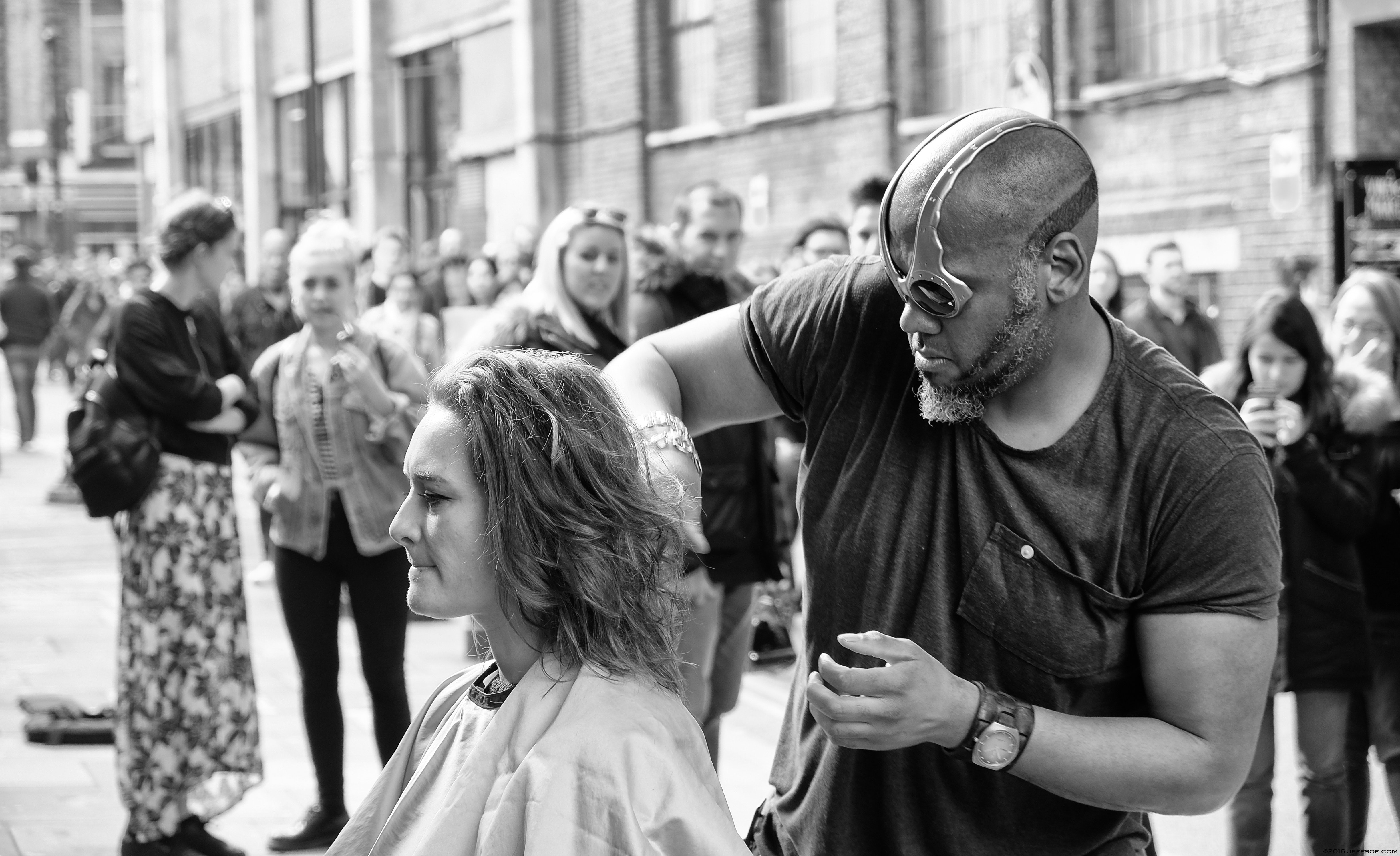 Woman having her hair cut by a man outside in a public space.